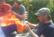 cozy chaos strickattacke im Schrebergarten Bochum. Drei Männer stricken an einem orangefarbenen Kunstobjekt, welches aus einem blauen Rollkoffer wächst.